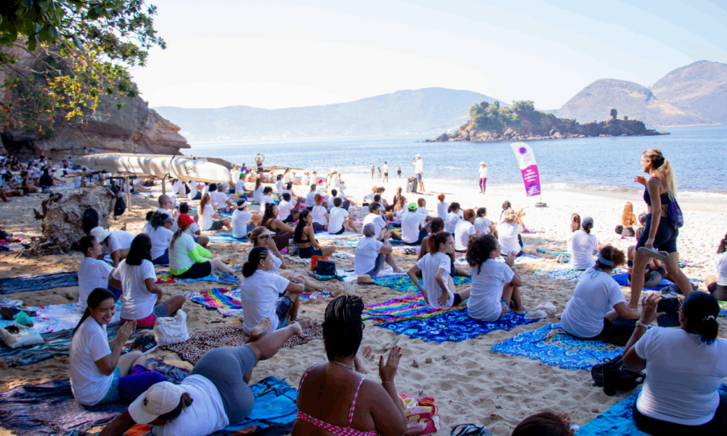 Grupo de pessoas praticando yoga sobre cangas na areia da Praia da Boa Viagem, em Niterói, com o Museu de Arte Contemporânea (MAC) ao fundo durante o amanhecer.