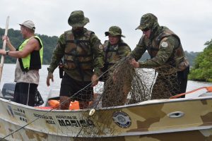 Agentes da Guarda Municipal de Maricá e da Secretaria de Pesca em barco patrulhando canal lagunar; ao fundo, vegetação de restinga e redes de pesca apreendidas.