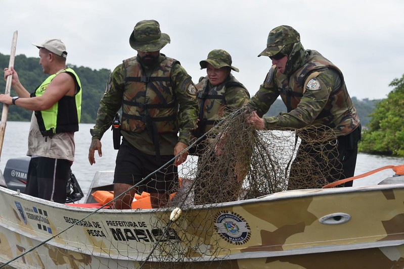 Agentes da Guarda Municipal de Maricá e da Secretaria de Pesca em barco patrulhando canal lagunar; ao fundo, vegetação de restinga e redes de pesca apreendidas.