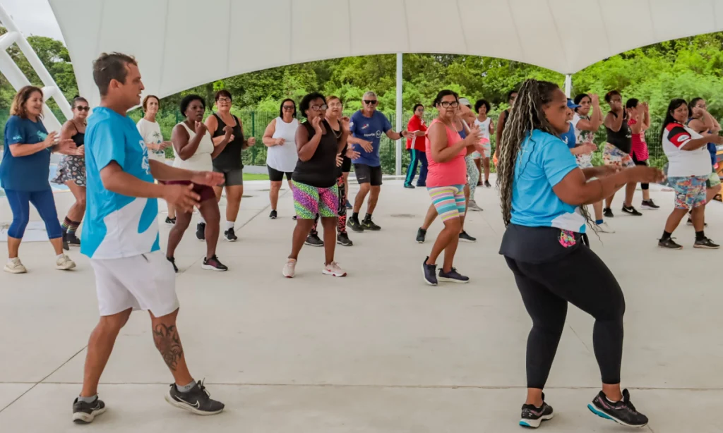 Grupo de pessoas praticando aula de dança ao ar livre na área de eventos do Parque RJ Nosso Sonho, no bairro Boa Vista, em São Gonçalo, sob a luz do dia.