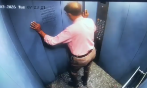 Câmera de segurança mostra o interior de um elevador metálico. Um homem de camisa rosa e calça escura tenta sair da cabine que está em movimento ascendente com a porta aberta, em um momento de perigo iminente.