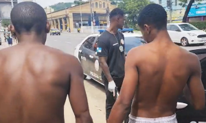 jovens de costas sem camisa, algemados, sendo conduzidos para a viatura policial
