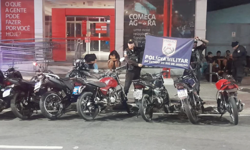 Motos aprendidas pela Polícia Militar, enfileiradas lado a lado na frentre de uma agência bancária, no Centro de São Gonçalo. Polícias mostram bandeira com símbolo do batalhão da cidade