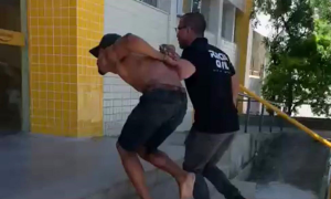 Homem algemado e sem camisa é conduzido por um policial ao interior de uma delegacia.