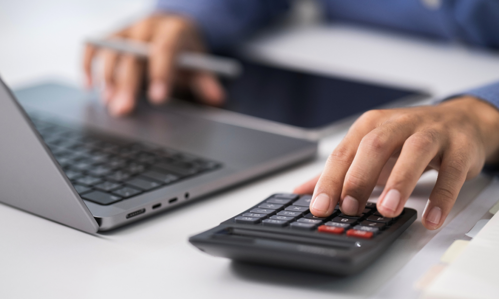 Close-up de uma mão operando uma calculadora e um computador ao lado. Sobre a mesa, documentos contábeis e uma calculadora sugerem organização financeira.