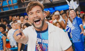 Jan Oliveira sorridente, vestindo camisa azul e branca da Portela, em frente ao barracão da escola com o símbolo da águia ao fundo.