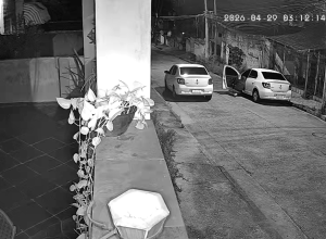 Imagem de câmera de segurança em preto e branco mostrando dois carros brancos estacionados lado a lado em uma rua residencial deserta durante a madrugada.