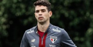 Oscar, com uniforme de treino do São Paulo, sorrindo durante entrevista no Centro de Treinamento de Cotia. Ao fundo, campos de futebol e o emblema do clube.